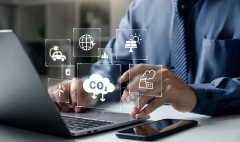 Eine Fachperson arbeitet an einem Laptop, umgeben von digitalen Symbolen für CO2-Emissionen, eine Fabrik, erneuerbare Energie und ein Elektrofahrzeug – sinnbildlich für die Antwort auf die Frage, was sind Scope 1 2 und 3 Emissionen im Fuhrpark-Reporting